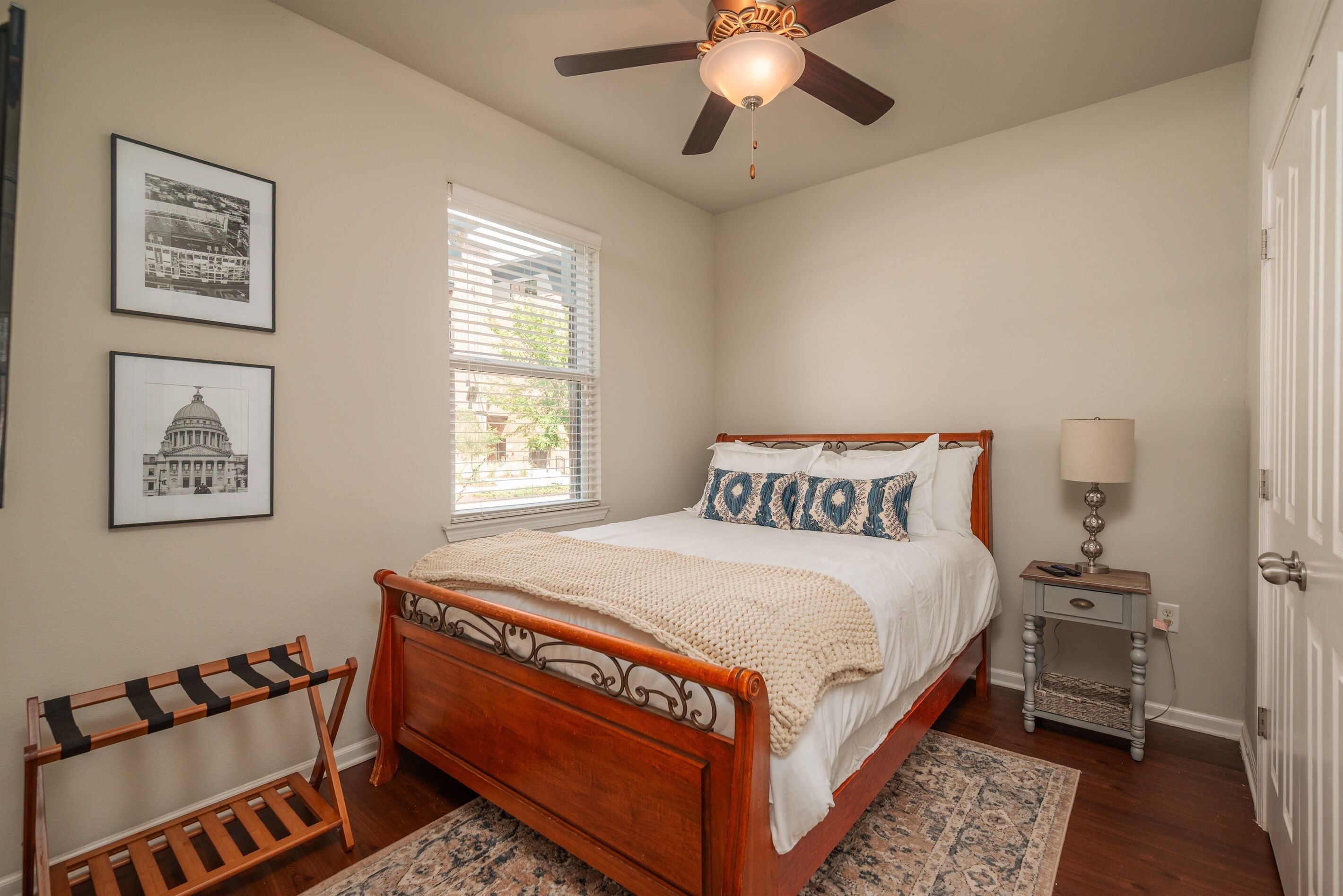 A bedroom with a wooden bed, a ceiling fan, a window with blinds, and two framed pictures on the wall.