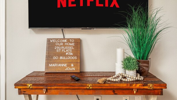 Un televisor pantalla plana fixat a la paret, amb un cartell de "NETFLIX" a sobre. Sota el televisor, hi ha un cartell de benvinguda de fusta amb un número de carrer i noms. Un comandament a distància reposa sobre una taula de fusta al costat del televisor, que també conté elements decoratius com una planta i un ramó de flors.