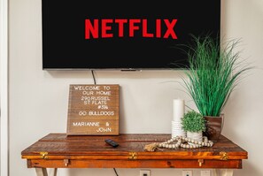 Un téléviseur à écran plat est fixé au mur, avec un panneau « NETFLIX » au-dessus. En dessous du téléviseur, on trouve un panneau en bois servant de panneau d’accueil, sur lequel figurent une adresse et des noms. Une télécommande est posée sur une table en bois à côté du téléviseur, qui contient également des objets décoratifs, comme une plante et un vase.