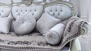 Um sofá branco luxuoso com almofadas acolchoadas e um cobertor estampado, posicionado diante de uma parede branca com um lustre moderno.