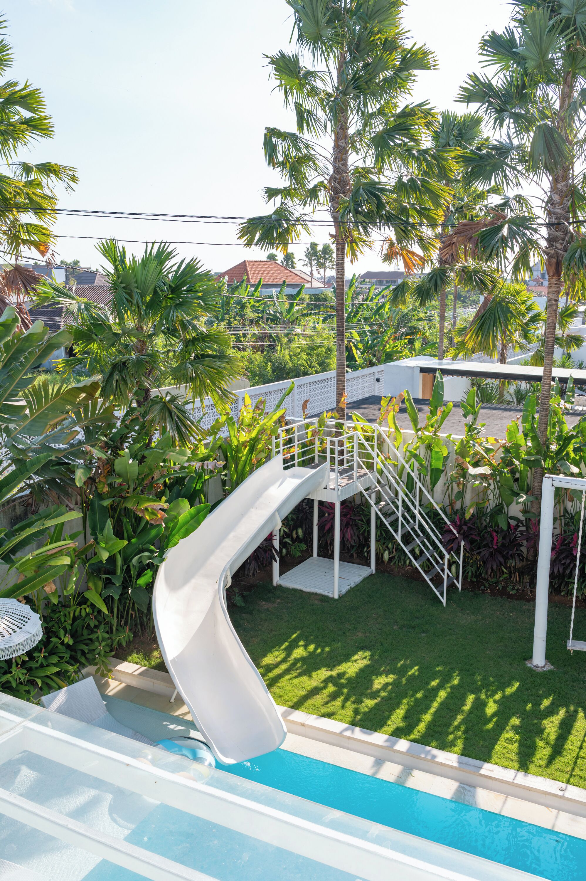 A pool with a slide, surrounded by palm trees and a white fence.