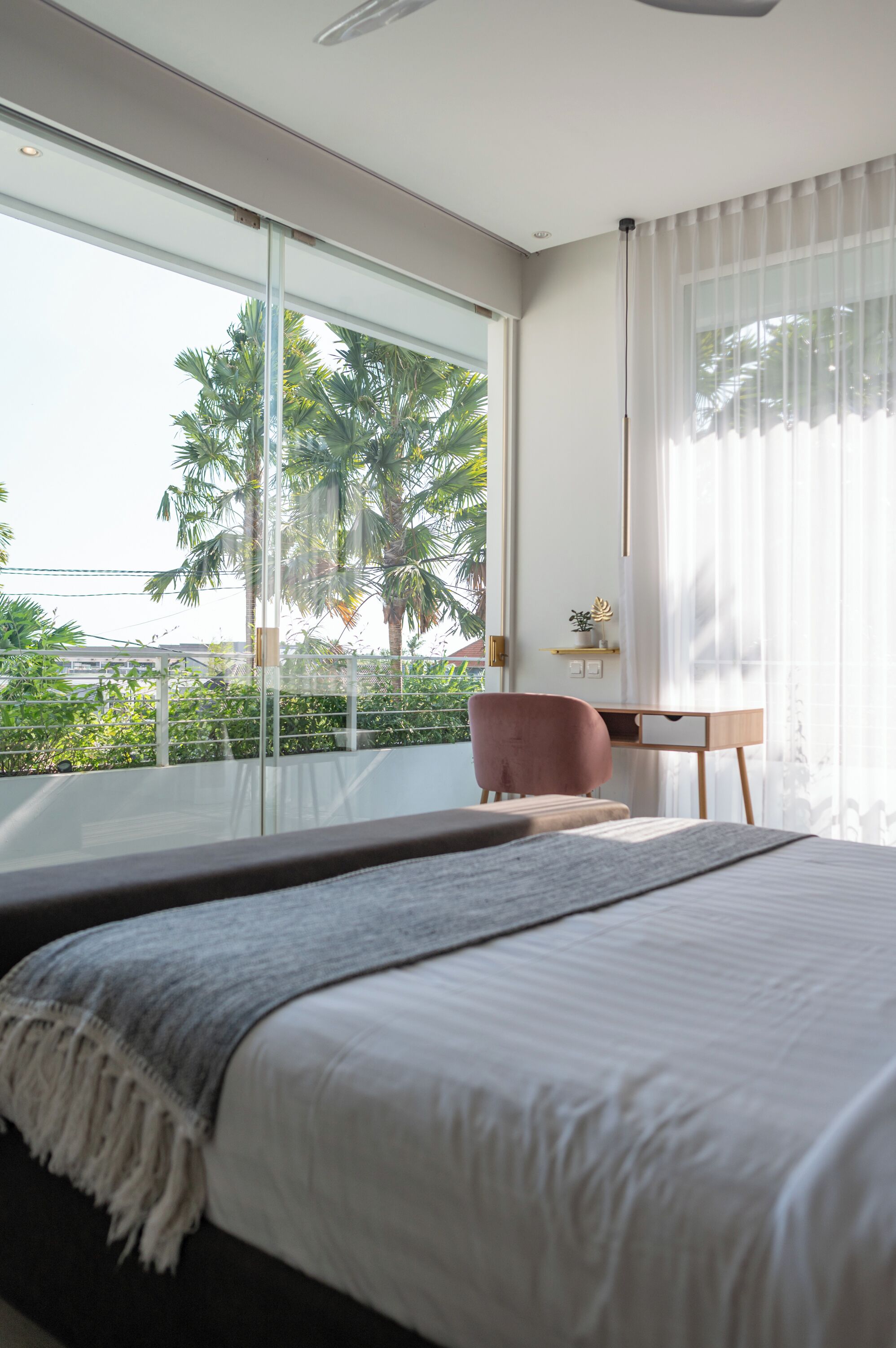 Un dormitorio con una cama grande, un escritorio y una silla. Se ve una ventana con vistas a palmeras y un balcón.