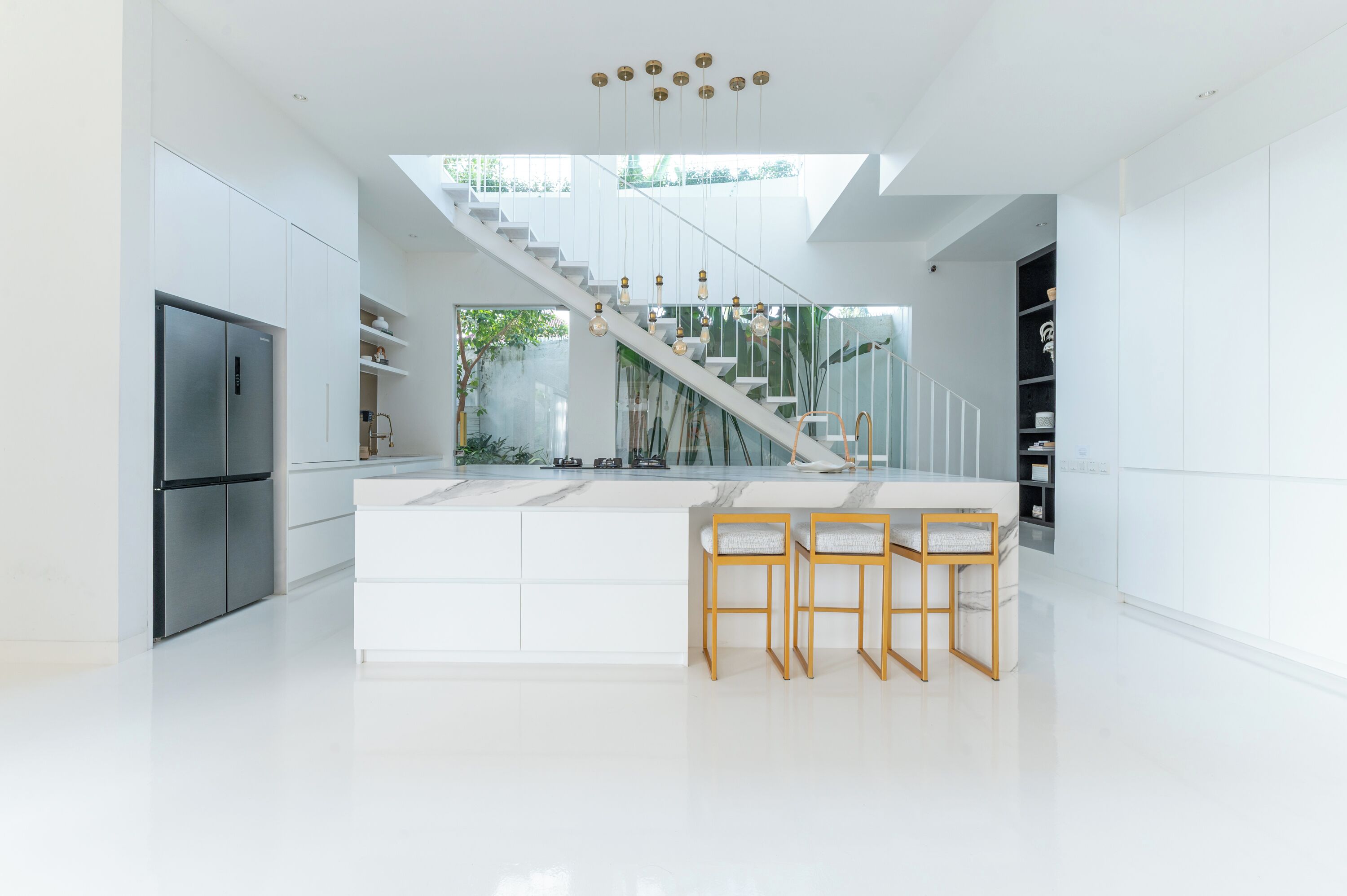 A modern kitchen with a central island, white cabinetry, a stainless steel refrigerator, and a staircase with glass railings.