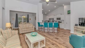 Een ruime woonkamer met een bank, een salontafel en een eethoek met barstoelen. In de achtergrond is een keuken met witte kasten en een plafondventilator zichtbaar.