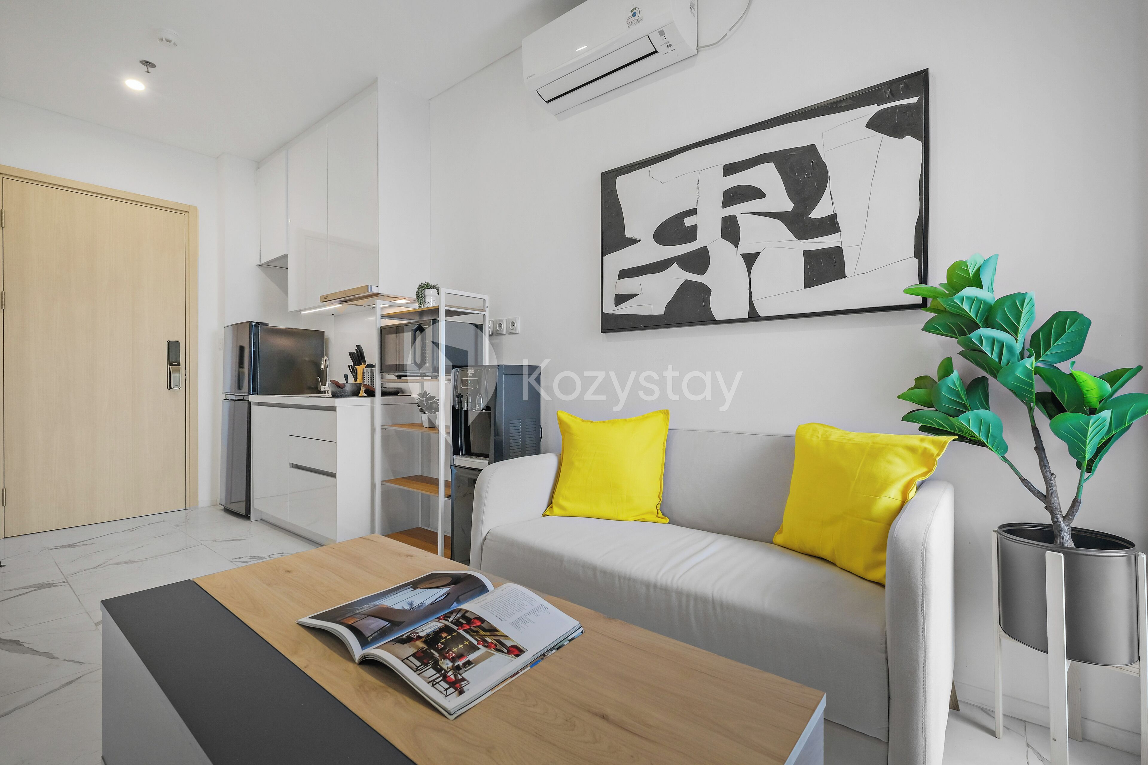 A modern living room with a sofa, a coffee table, and a magazine. There is a kitchen area with a refrigerator and a microwave.
