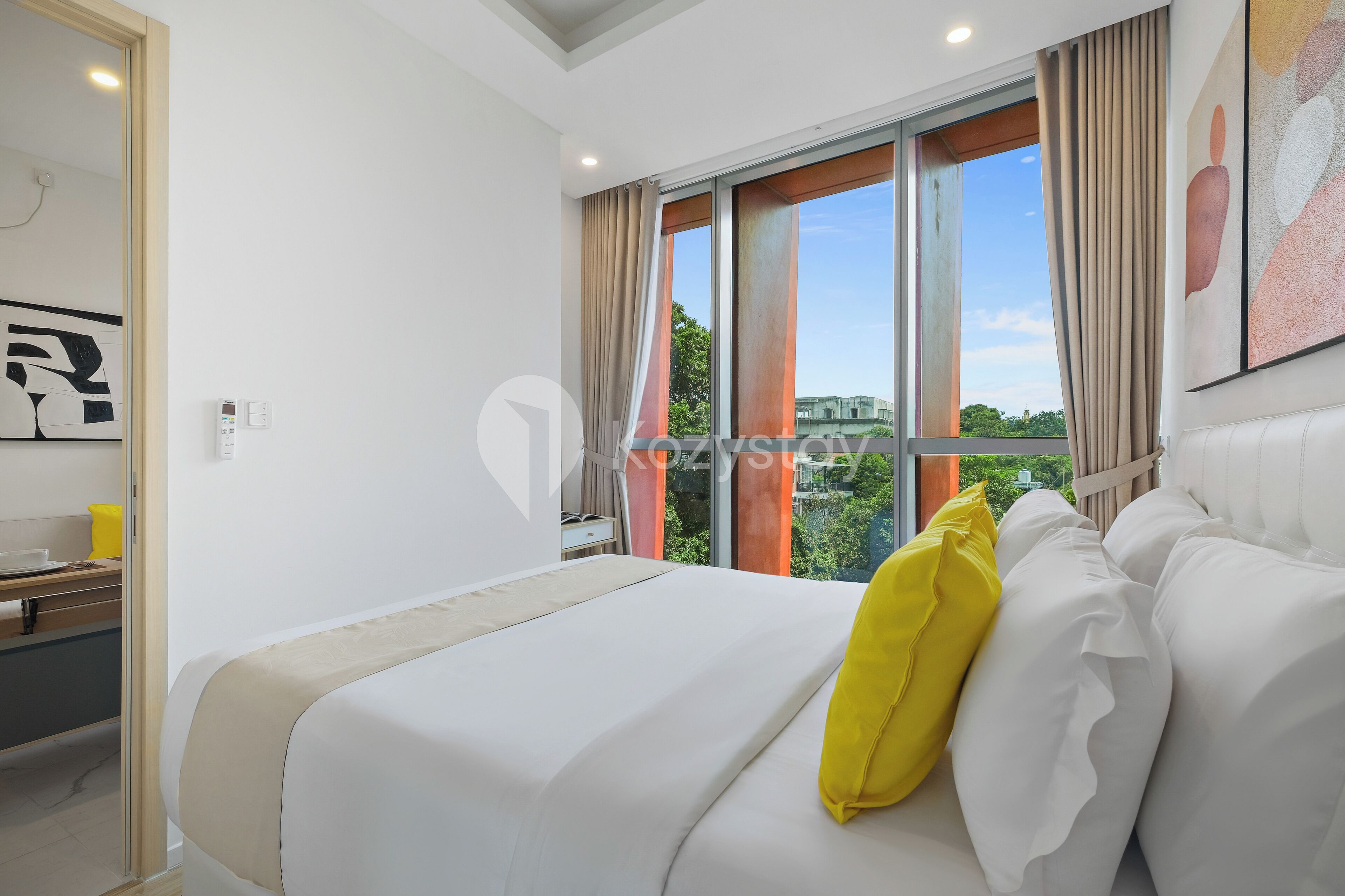 A hotel room with a bed, a window with curtains, a painting on the wall, and a view of greenery outside.