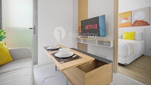Ein modernes Hotelzimmer mit einem Holztisch, einem an der Wand befestigten Fernseher und einem Bett mit einem gelben Kopfkissen.