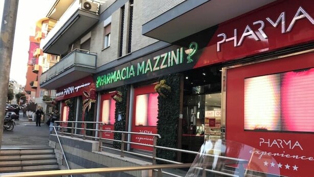 Vista da rua com uma farmácia chamada 'PHARMACIA MAZZINI' e um prédio com fachada vermelha.
