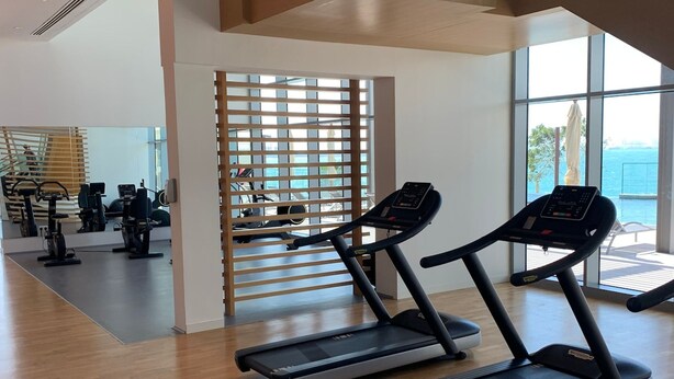 Ein Fitnessraum mit Laufbändern, Ergometer und einer Treppe, die zu einem Balkon mit Aussicht führt.
