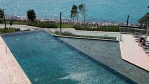 Ein rechteckiger Swimmingpool mit klarem, blauem Wasser, umgeben von einer gepflasterten Liegefläche. Im Hintergrund Blick auf das Meer und den fernen Stadthintergrund.