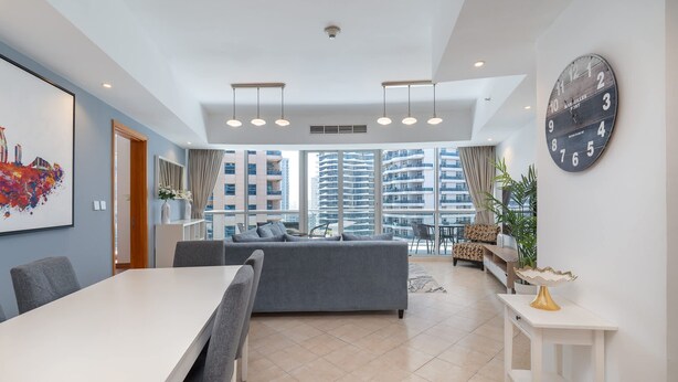 A modern living room with a grey sofa, a dining table, and a large wall clock. - Spacious 3BR Marina Stay With City Views (Dubai)