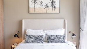 A bedroom with a large bed, two black and white patterned pillows, a wall-mounted painting of palm trees, and bedside lamps. - Chic 1-bed for 4 in Prime Dubai Hills Location (Dubai)