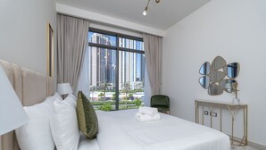 A hotel room with a large bed, a view of tall buildings, a green chair, and a decorative mirror on a console table. - Bright Elegant 2BR in Creek Harbour (Dubai)