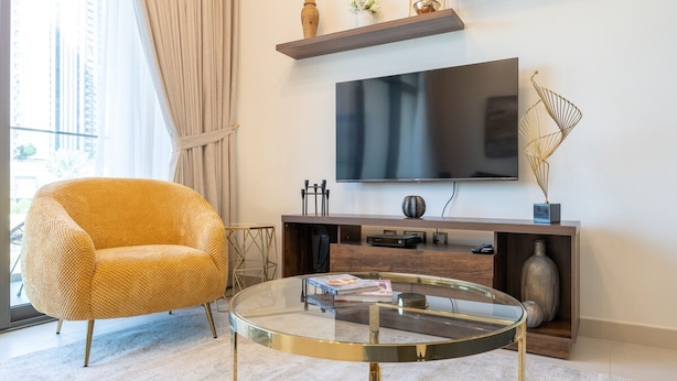 A modern living room with a round glass coffee table, a yellow armchair, a flat-screen TV mounted on a wooden console, and a decorative metal sculpture. - Bright Elegant 2BR in Creek Harbour (Dubai)