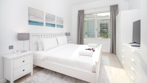 Une chambre à coucher moderne avec un lit blanc, des tables de chevet, un téléviseur et des œuvres d’art accrochées au mur.