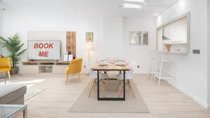 Un salon moderne avec une table à manger en bois, disposée pour quatre personnes, des chaises blanches, un téléviseur fixé à un mur blanc, une bibliothèque et une plante en pot.