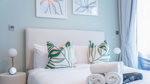 A neatly made bed with white bedding and green leaf-patterned pillows, a bedside lamp, and a rolled towel on a bedside table. - Glamorous 1BR Apartment - Steps From the Beach (Dubai)