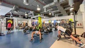 Une salle de sport équipée de divers appareils d’exercice, avec un sol bleu, et une personne utilisant une machine à presses pour les jambes.