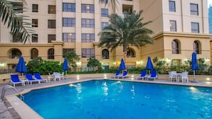 Piscina con sillas y sombrillas azules, rodeada de un edificio con múltiples ventanas y palmeras.