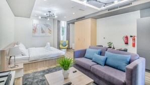 Una habitación de hotel moderna con cama, sofá y una pequeña mesa con una planta. Hay una ventana con cortinas, una lámpara en el techo y un extintor en la pared.