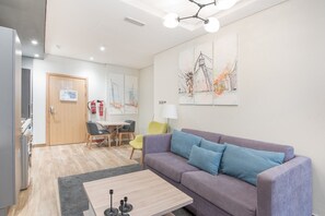 Een moderne woonkamer met een bank, een salontafel, en een eetgedeelte met stoelen. Er is een deur met een bordje, een brandblusser, en kunst aan de muur.