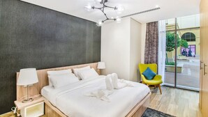 Une chambre d’hôtel moderne avec un grand lit, une table de chevet avec une lampe, un lustre lumineux, un fauteuil jaune et une vue sur l’extérieur grâce à une porte vitrée.