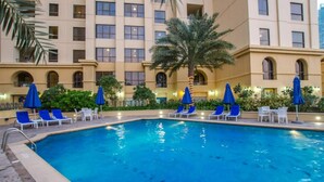 A swimming pool with blue chairs and umbrellas, surrounded by greenery and a multi-story building with arched windows. - Steps by the Beach - Serene Studio in JBR (Dubai)