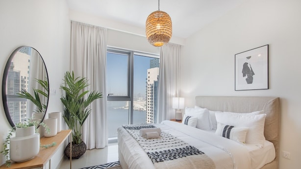 A modern hotel room with a large bed, a view of the city, a hanging light fixture, and a potted plant. - Stylish Cozy 3-bedroom Apartment With Harbour Views in Dubai Creek Harbour (Dubai)