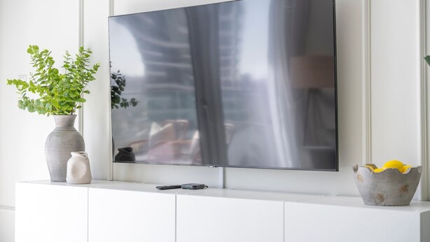 Ein Flachbildfernseher an einer weißen Wand, darunter eine weiße Ablagefläche mit einem Blumentopf und grünen Pflanzen sowie einer Schale gelber Zitronen.