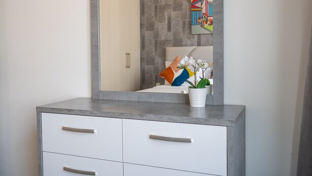 A bathroom with a large mirror, a white cabinet with drawers, a potted orchid, and a colorful painting on the wall. - Chic 1BR Flat Stunning Views in Dubai Hills (Dubai)
