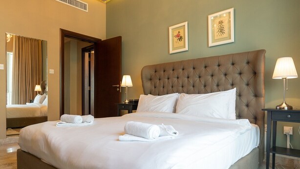 A neatly made bed with white linens and rolled towels, a tufted headboard, and two framed artworks on the wall. - Amazing 4-bedroom Apartment Perfect for Families in Dubai Marina (Dubai)