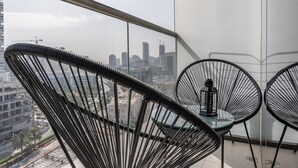 Un balcon avec deux chaises à l’assise en fil de fer noir et une petite table, d’où l’on peut admirer un paysage urbain avec des immeubles et des grues de construction.