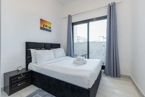 A modern bedroom with a large bed, a nightstand, a painting, and a view through a window. - Breathtaking 2BR Apartment in Jebel Ali Village (Dubai)