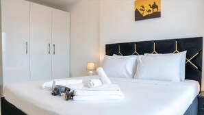 Ein ordentlich bezogenes Bett mit weißen Leinen und Handtüchern, ein schwarzes Kopfteil mit goldenen Kleiderbügeln sowie ein an der Wand hängendes Kunstwerk, das Kamele zeigt.
