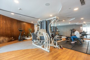 A modern gym with various exercise equipment, a wooden wall, and a person using a bench press. - Lovely 1-bedroom Apartment Perfect for 4 in JLT (Dubai)