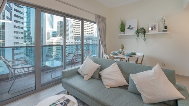 A modern living room with a grey sofa, white cushions, a dining table with chairs, and a balcony with a view of tall buildings. - Classy 1-bedroom Apartment in Dubai Marina With City Views (Dubai)