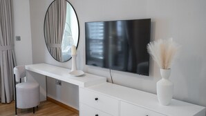 Une chambre à coucher moderne avec une coiffeuse blanche, un miroir rond, un téléviseur à écran plat et un vase contenant des plantes séchées.