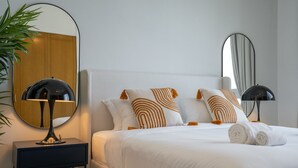 Un lit bien fait, avec une literie blanche et des coussins à motifs, un miroir rond accroché au mur et une petite table avec une lampe à côté.