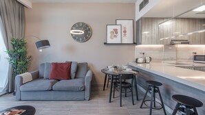 A modern living room with a grey sofa, a coffee table, a dining area with stools, a kitchen with a counter, and a wall clock. - Warm 1-bedroom Family-friendly Aparment in JLT (Dubai)