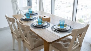 A dining table set with blue glasses, plates, and cutlery, surrounded by white cushioned chairs, in a room with a large window. - Elegant 2-bedroom Apartment in Dubai South Close to Expo 2020 (Dubai)
