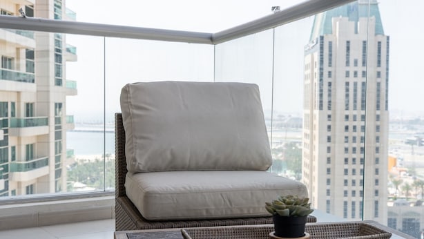 A balcony with a cushioned chair, a small table with a plant, and a view of a tall building and a body of water. - Bright 1-bedroom With View Dubai Marina (Dubai)