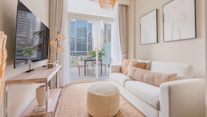 A modern living room with a large flat-screen TV, a white sofa, a round ottoman, and a balcony with a city view. - Bali-inspired 1-bedroom Apartment for 4 in Business Bay (Dubai)