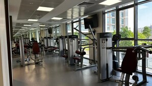 A gym with various exercise machines, large windows, and a flat ceiling with recessed lighting. - Stylish Homey 1-bedroom Apartment Perfect for 4 in Dubai Hills (Dubai)