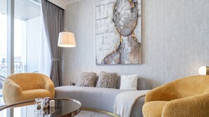 Een moderne hotelkamer met een grijze bank, twee gele fauteuils, een glazen salontafel en een groot abstract schilderij aan de muur.