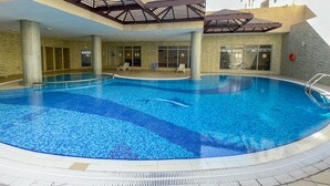 Una piscina interior con suelo de baldosas, rodeada por un edificio con varias ventanas y una zona techada sobre la piscina.