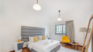 Un dormitorio moderno con una cama tapizada en gris, ropa de cama blanca, una silla amarilla y un ventanal con vistas a edificios.