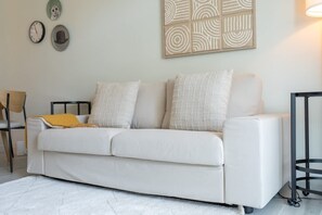 Un canapé blanc agrémenté de plusieurs coussins, une petite table avec une lampe et des œuvres d’art murales, notamment une horloge et des motifs abstraits.
