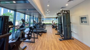A gym with cardio machines, weightlifting equipment, and a large window offering a view of the outdoors. - Stylish 1-bedroom Apartment With Stunning Canal Views at Business Bay (Dubai)