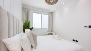 A modern bedroom with a large bed, white bedding, a potted plant, and a view of the city through the window. - Elegant 1-bedroom Apartment in Jumeirah Village Triangle With Balcony Bliss (Dubai)