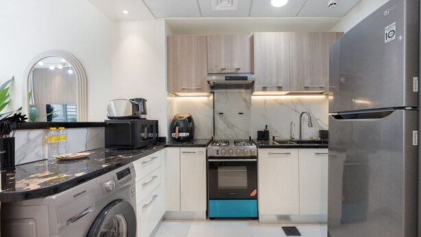 A modern kitchen with a washing machine, a microwave, a stove, and a refrigerator. - Elegant 1-bedroom Apartment in Jumeirah Village Triangle With Balcony Bliss (Dubai)